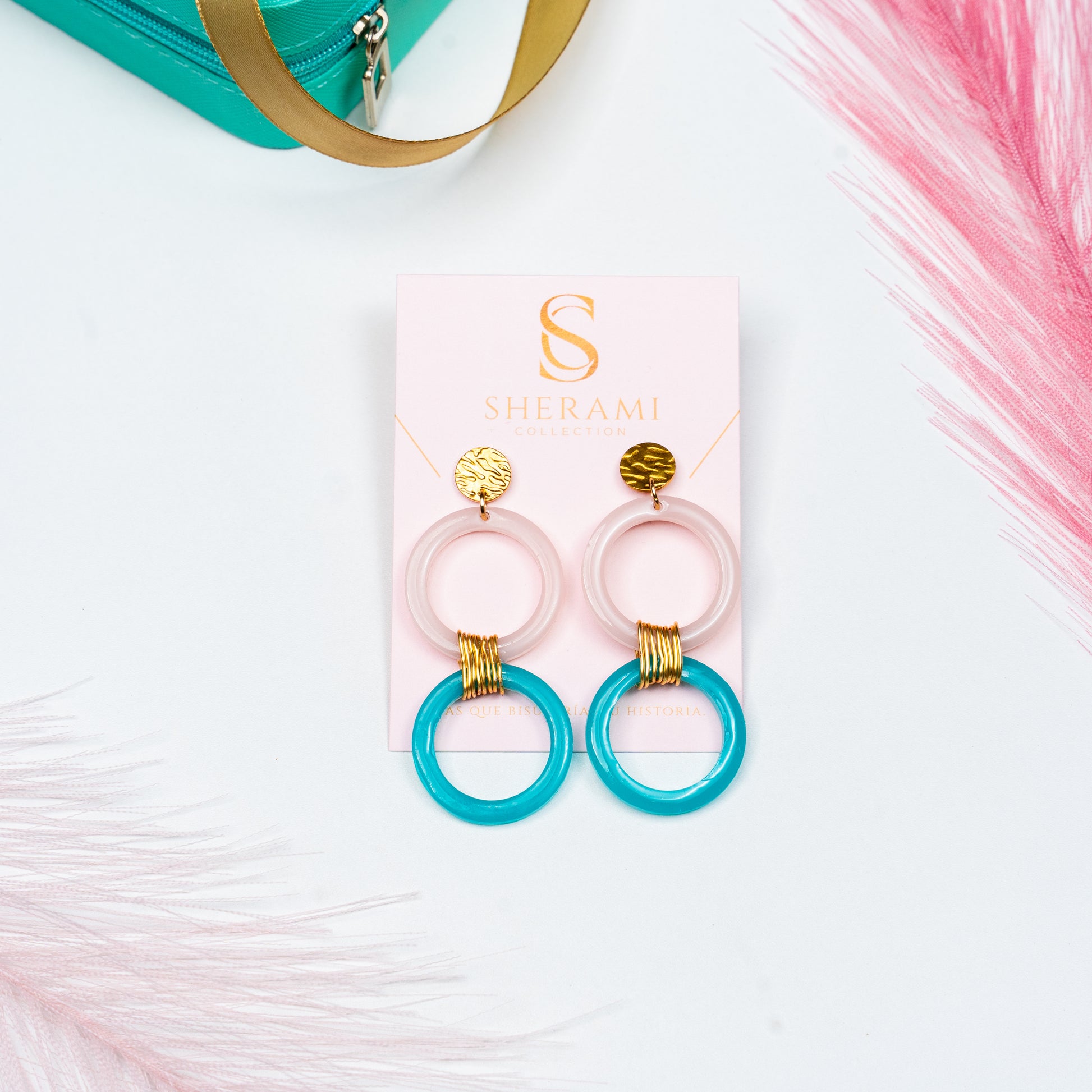 Par de aretes rosa, azul y dorado sobre la tarjeta de presentacion de Sherami Collection en un fondo blanco