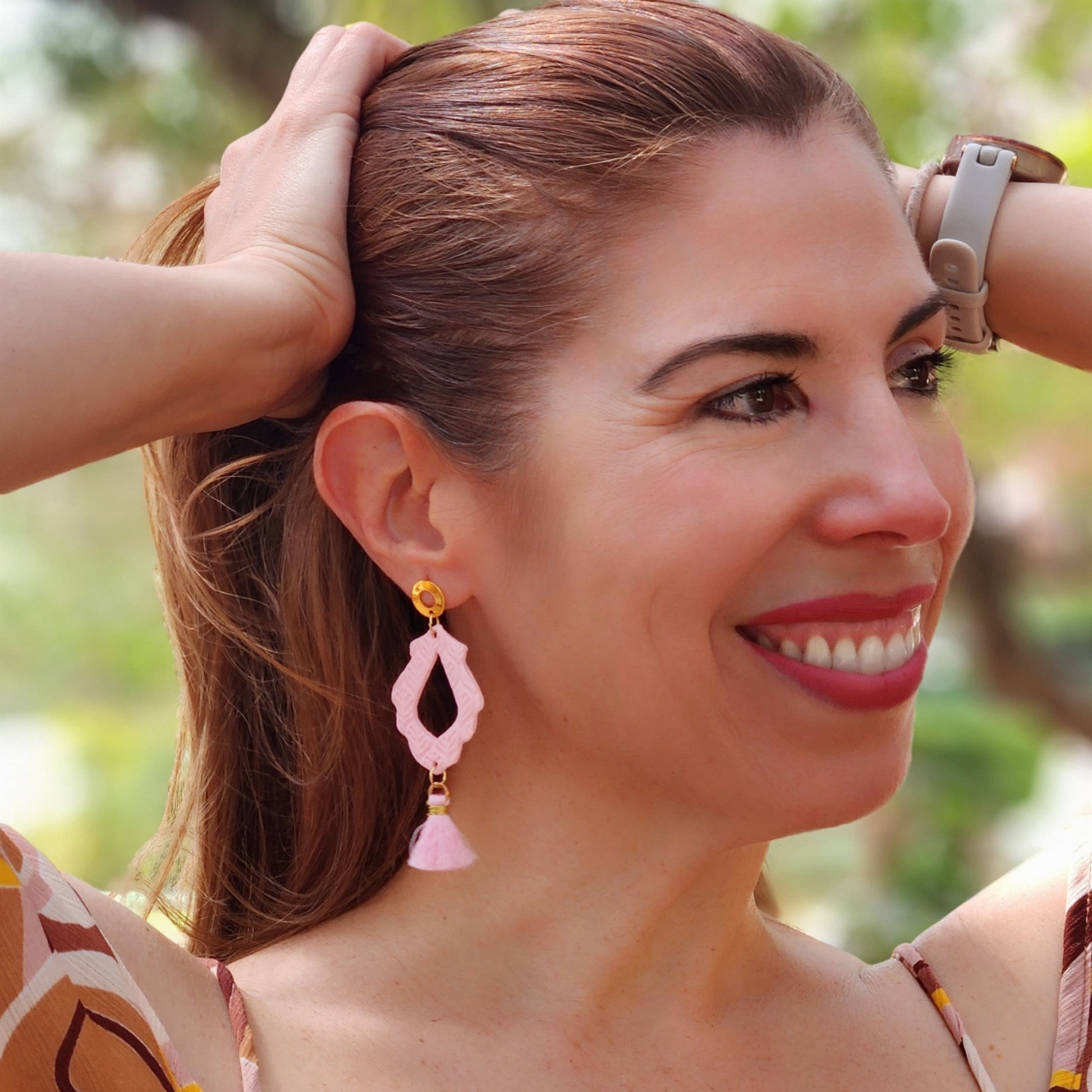 Woman wearing pink earrings with a blurred green background. Aretes artesanales Sherami Collection