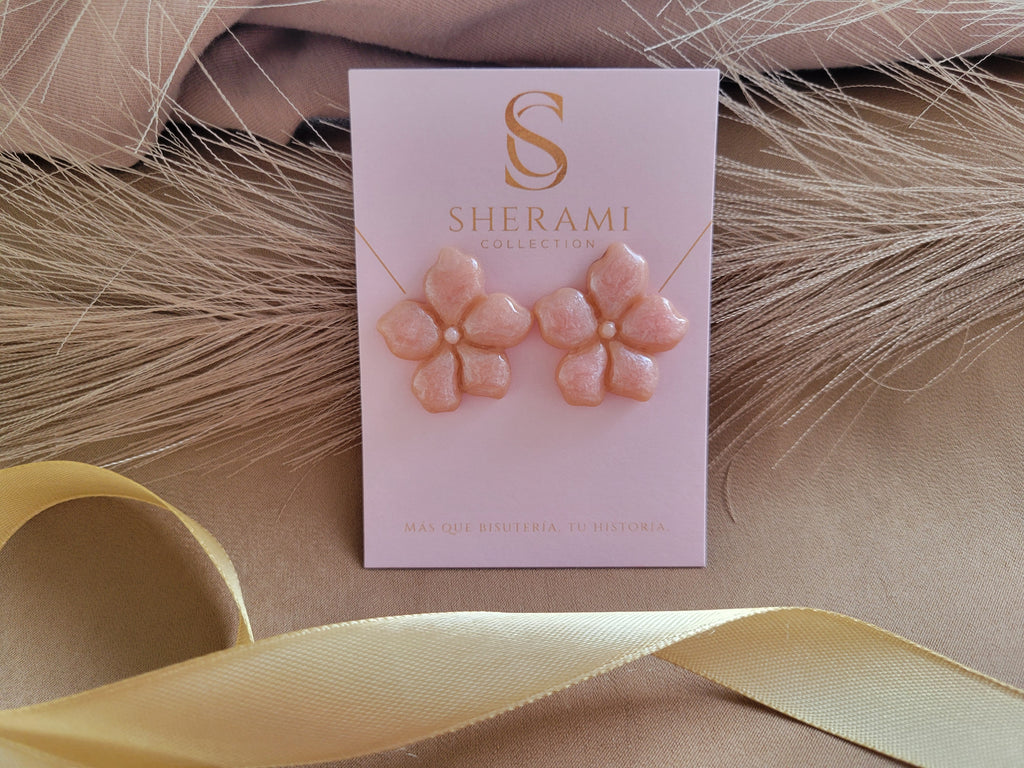 Pink flower-shaped earrings on a Sherami branded card with ribbons in the background. Aretes artesanales