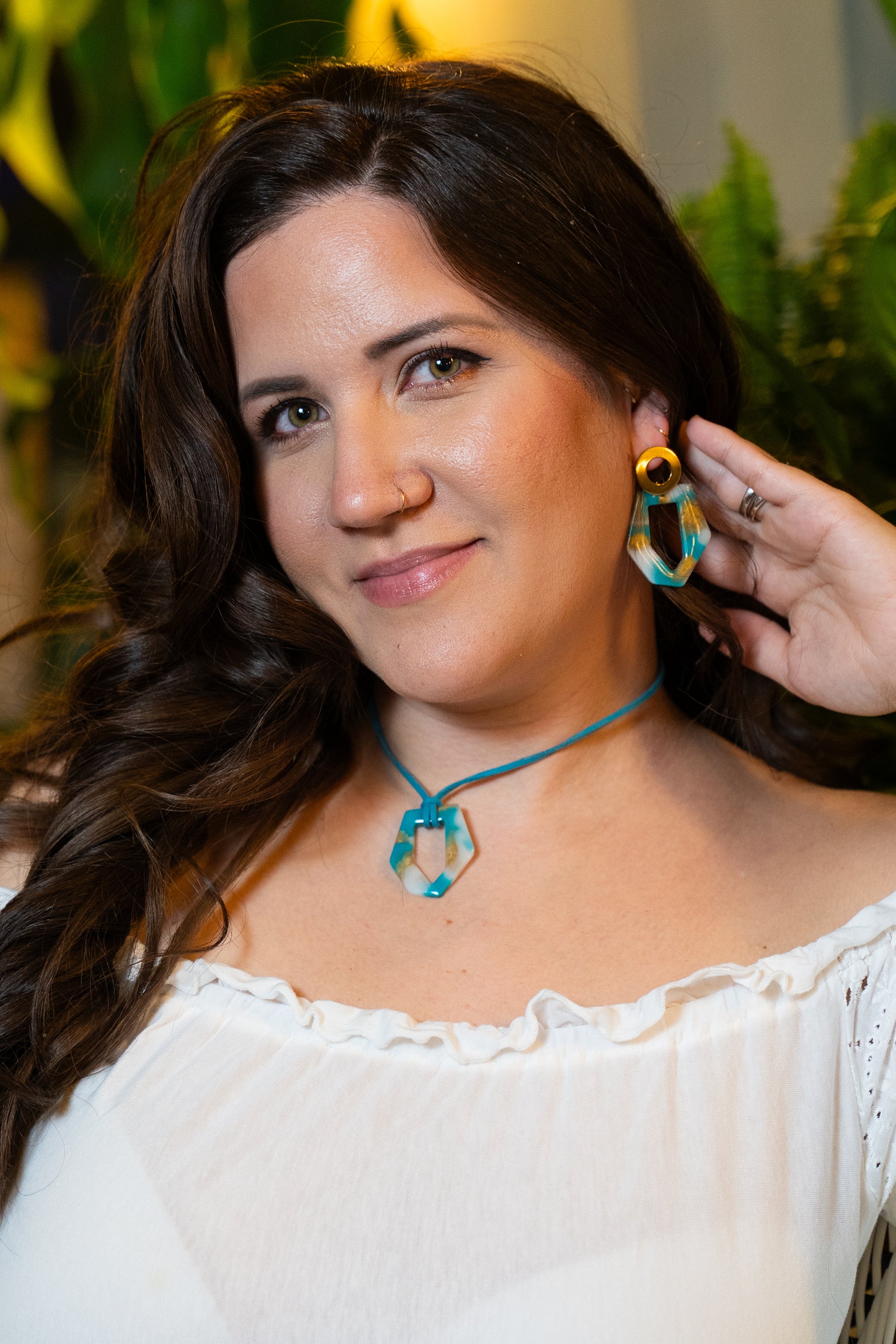 Mujer con aretes atesanales azul, blanco y dorado y collar a juego con un fondo verde borroso de Sherami Collection