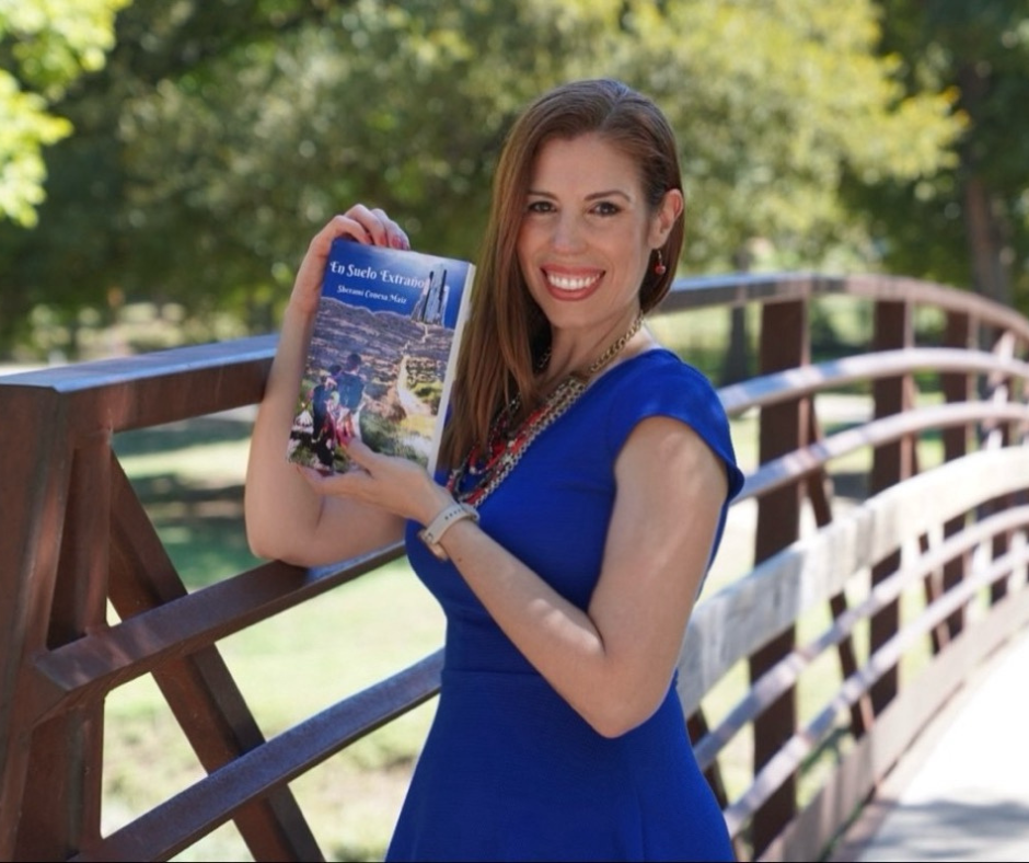Sherami Conesa con un vestido azul sosteniendo el libro En suelo extraño en un puente de madera con vegetación al fondo
