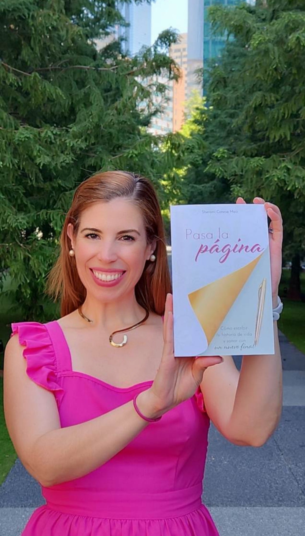 Sherami Conesa con un vestido rosa sosteniendo un libro titulado 'Pasa la Página' al aire libre con árboles y edificios en el fondo.