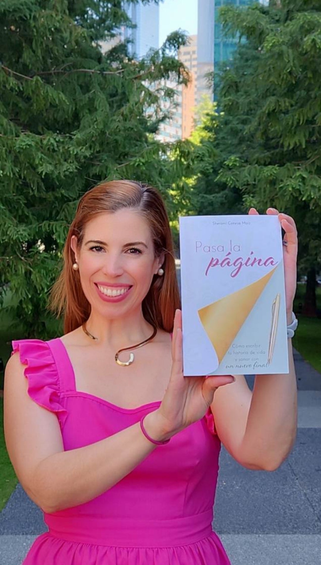 Sherami Conesa con un vestido rosa sosteniendo un libro titulado 'Pasa la Página' al aire libre con árboles y edificios en el fondo.