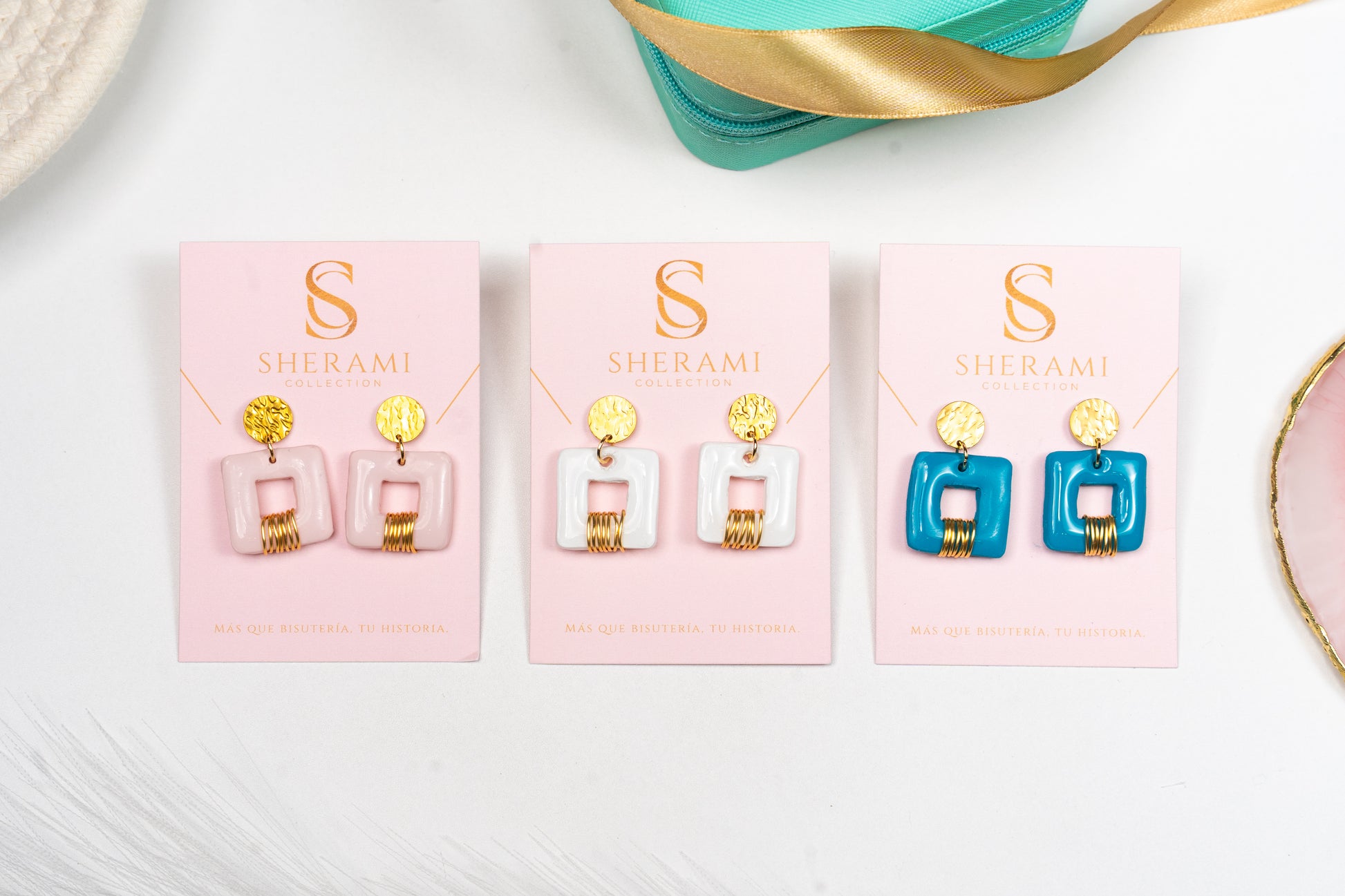 Tres pares de pendientes  modelo Invicta en exhibición con la marca 'Sherami Collection en tarjetas rosas.