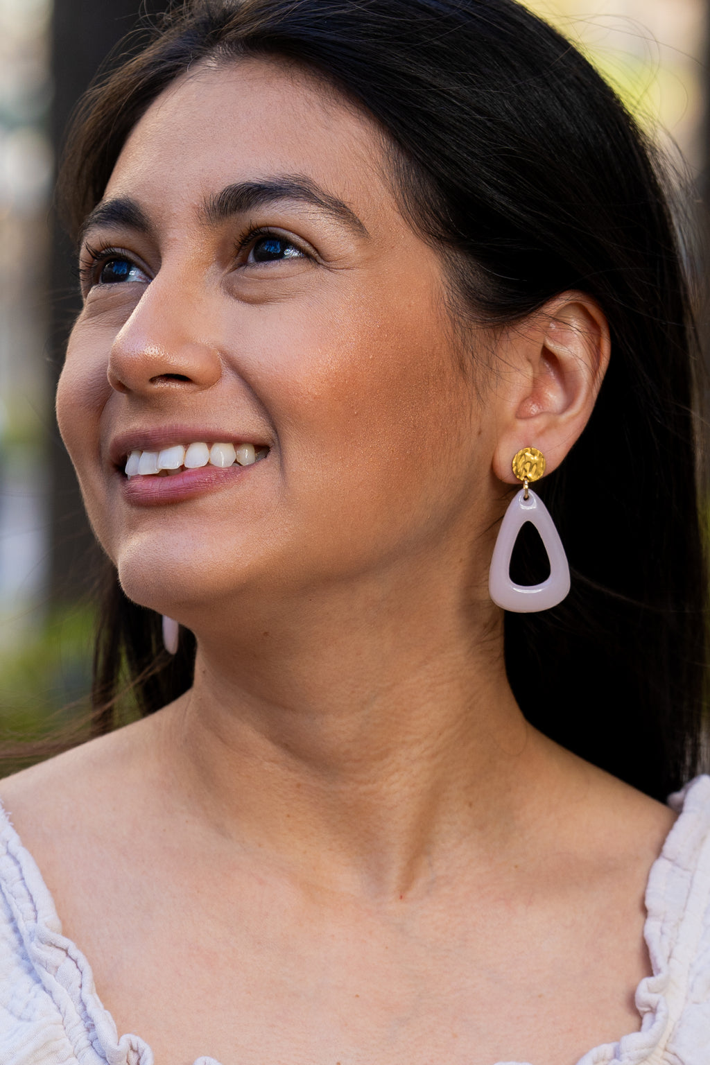 Woman wearing a white dress and light pink Sherami Collection earrings with a blurred outdoor background
