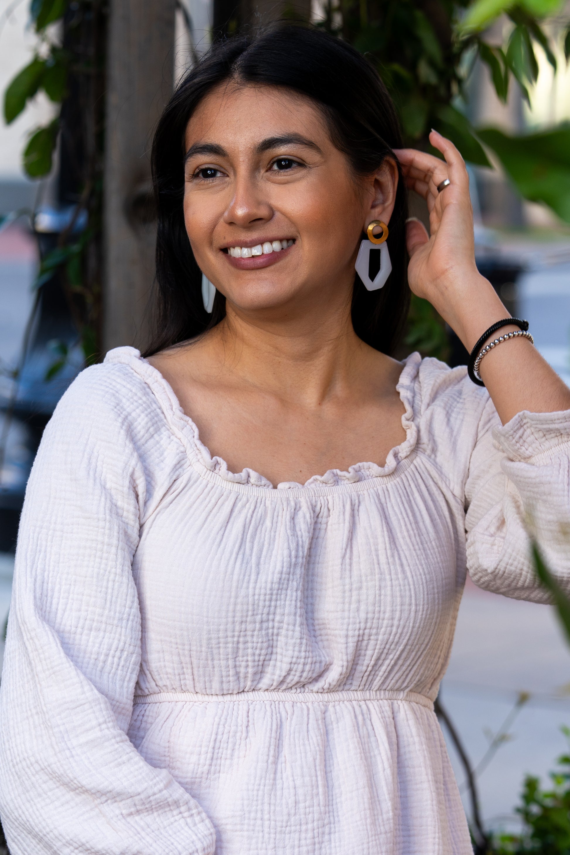 Woman in a white dress with a blurred background wearing Sherami Collection earrings