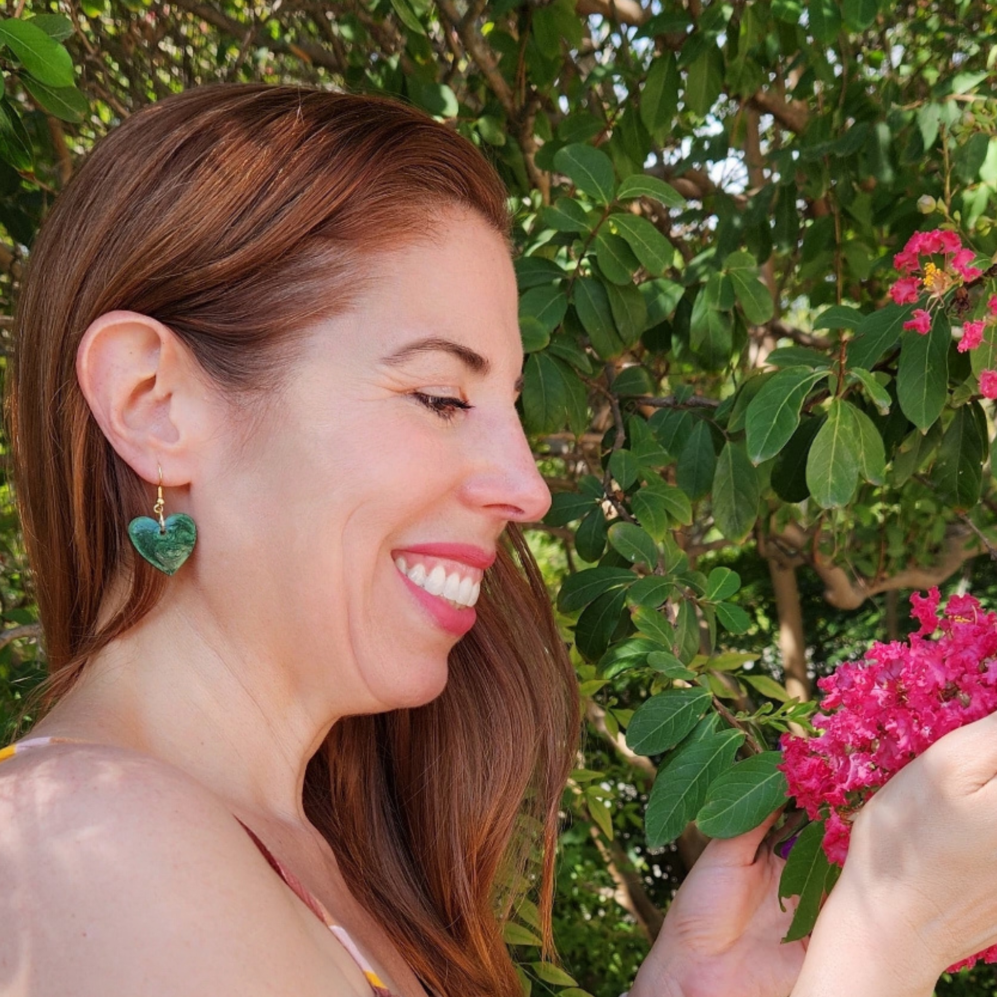 Woman with heart-shaped earrings smiling outdoors among greenery and pink flowers from Sherami Collection. joyeria artesanal hecha a mano